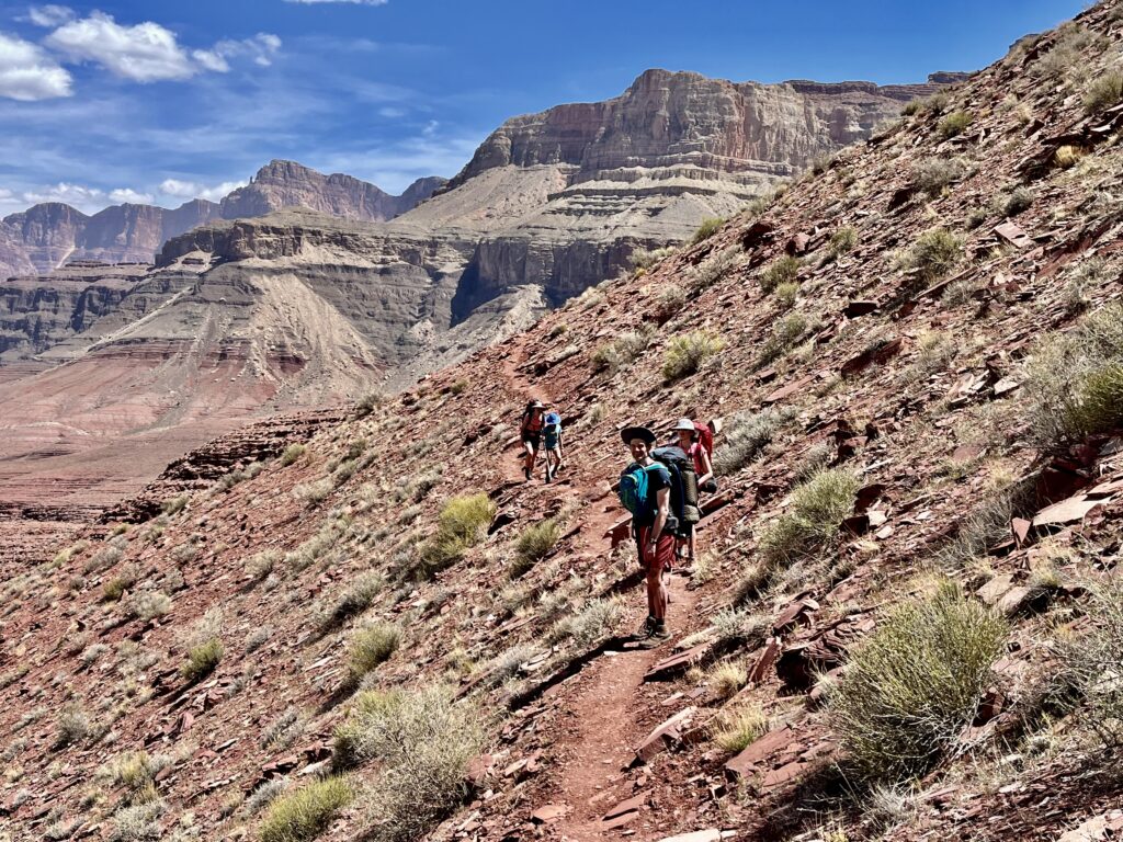 Group hikes a windswept trail with the Grand Canyon in the background.