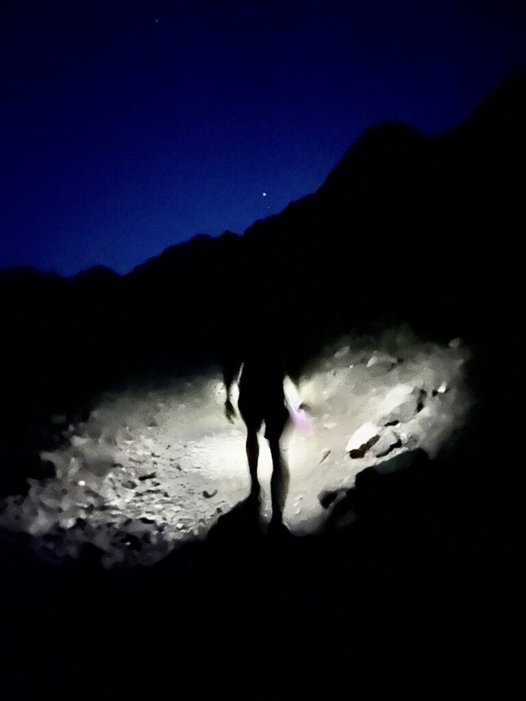 A headlamp brightens the trail in front of a silhouetted hiker.