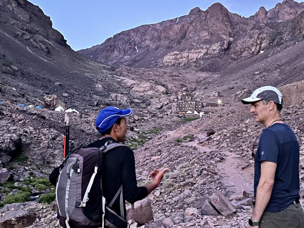 A guide and climber talk with a mountain backdrop including a large refuge.