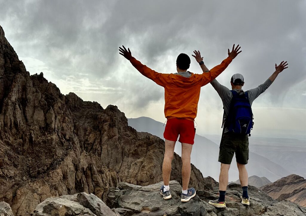 To men spread their arms above their heads, looking at a rocky mountain ridge.